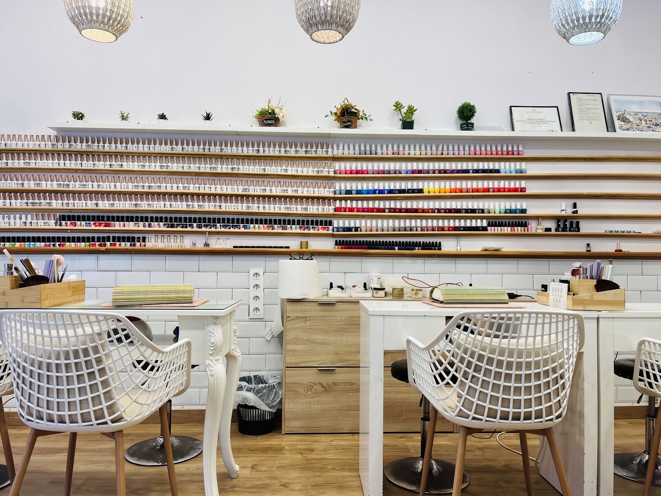 Interior del salón Fuego Nails: sillas blancas de malla, mesas de trabajo de madera y una pared llena de frascos de esmalte.